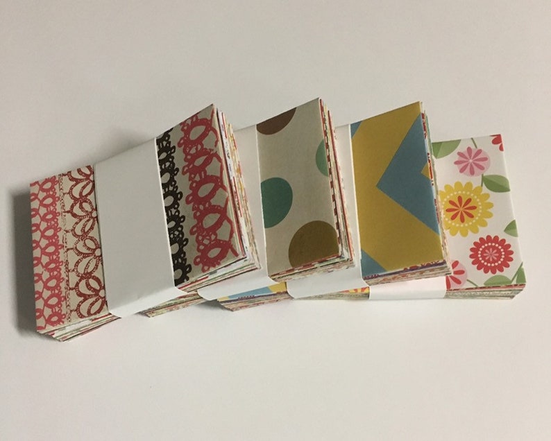Peut inclure: Quatre piles de carr&eacute;s de papier &agrave; motifs color&eacute;s, chaque pile &eacute;tant envelopp&eacute;e de papier blanc. Les motifs comprennent de la dentelle rouge et blanche, des pois bruns et blancs, des formes g&eacute;om&eacute;triques jaunes et bleues et des motifs floraux roses et jaunes.