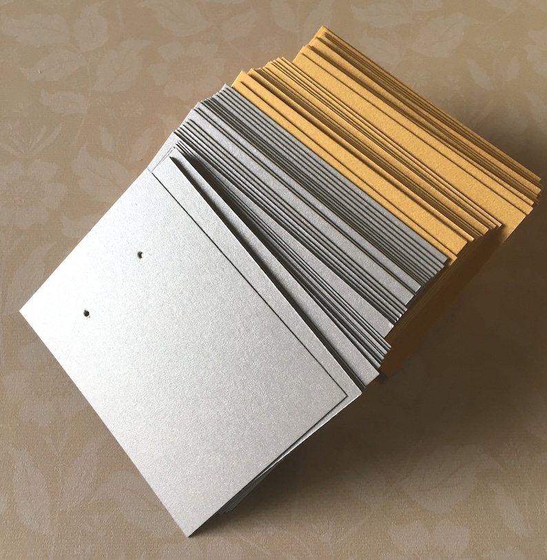 May include: A stack of earring display cards in silver and gold. The cards have a textured finish. Each silver card has two small holes. The cards are stacked at an angle, revealing the edges of the cards.
