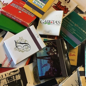 May include: A collection of vintage matchbooks with various designs and text. Some of the matchbooks feature images of hotels, restaurants, and businesses. The matchbooks are in various colors, including red, green, yellow, and blue.