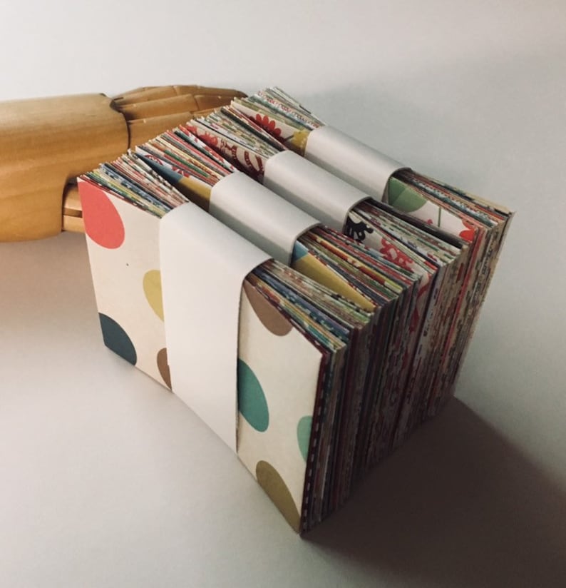 Peut inclure: Trois piles de papier &agrave; motifs color&eacute;s, chacune envelopp&eacute;e de papier blanc avec une bande blanche au milieu. Le papier est dispos&eacute; en quinconce.