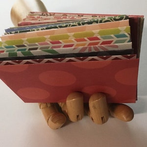 Peut inclure: Une pile d'enveloppes en papier &agrave; motifs color&eacute;s, tenue dans une main en bois. L'enveloppe du dessus est rouge avec des pois blancs.