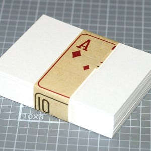 May include: A stack of blank white cards, bound with a band featuring a playing card design. The band displays the Ace of Diamonds and the number 10. The cards are on a gray grid surface.