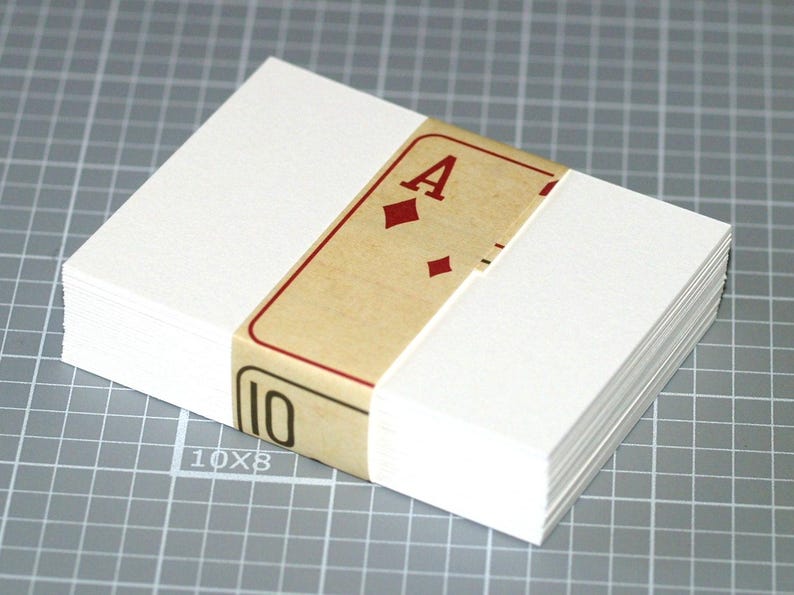May include: A stack of blank white cards, bound with a band featuring a playing card design. The band displays the Ace of Diamonds and the number 10. The cards are on a gray grid surface.
