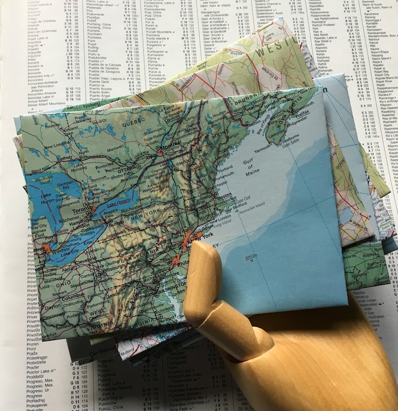 May include: A close-up of a wooden hand holding a stack of folded maps. The maps are of the United States and Canada, with a blue and green color scheme. The maps are folded and show the major cities and geographical features of the regions.