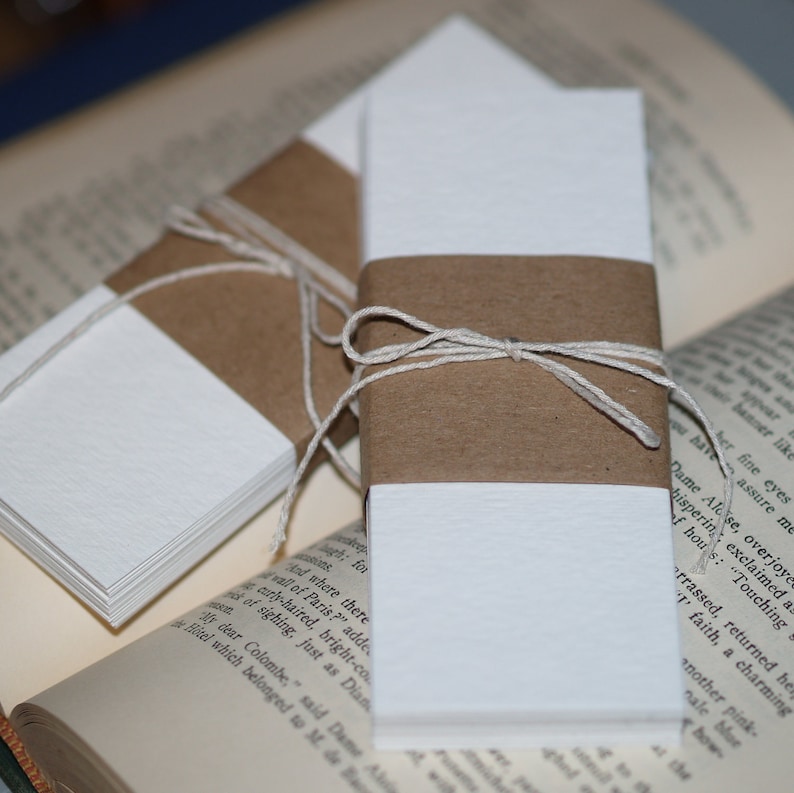 K&ouml;nnte beinhalten: Drei Stapel wei&szlig;er Papiernotizb&uuml;cher, die jeweils mit Schnur gebunden und in braunes Papier eingewickelt sind. Die Notizb&uuml;cher liegen auf einem Buch.