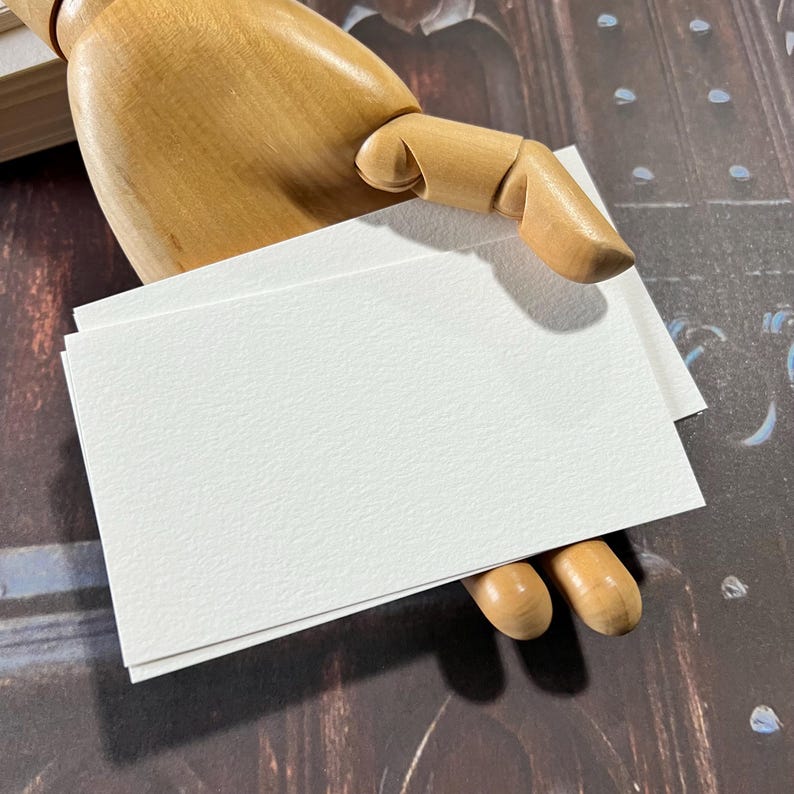 May include: A stack of blank, white rectangular cards with a textured surface. The cards are held by a wooden articulated hand. The background is a dark, patterned surface.