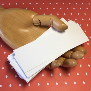 K&ouml;nnte beinhalten: Ein Stapel leerer, wei&szlig;er rechteckiger Karten, die von einer Holzhand gehalten werden. Die Karten sind ordentlich gestapelt, und die Holzhand befindet sich vor einem roten Hintergrund mit einem wei&szlig;en Tropfenmuster.