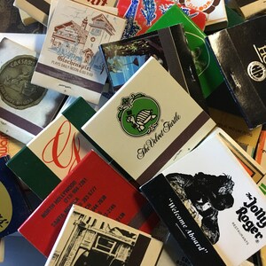 May include: A collection of vintage matchbook covers with various designs and colors. Some of the matchbooks feature images of buildings, animals, and text. The matchbooks are arranged in a pile, with some overlapping.
