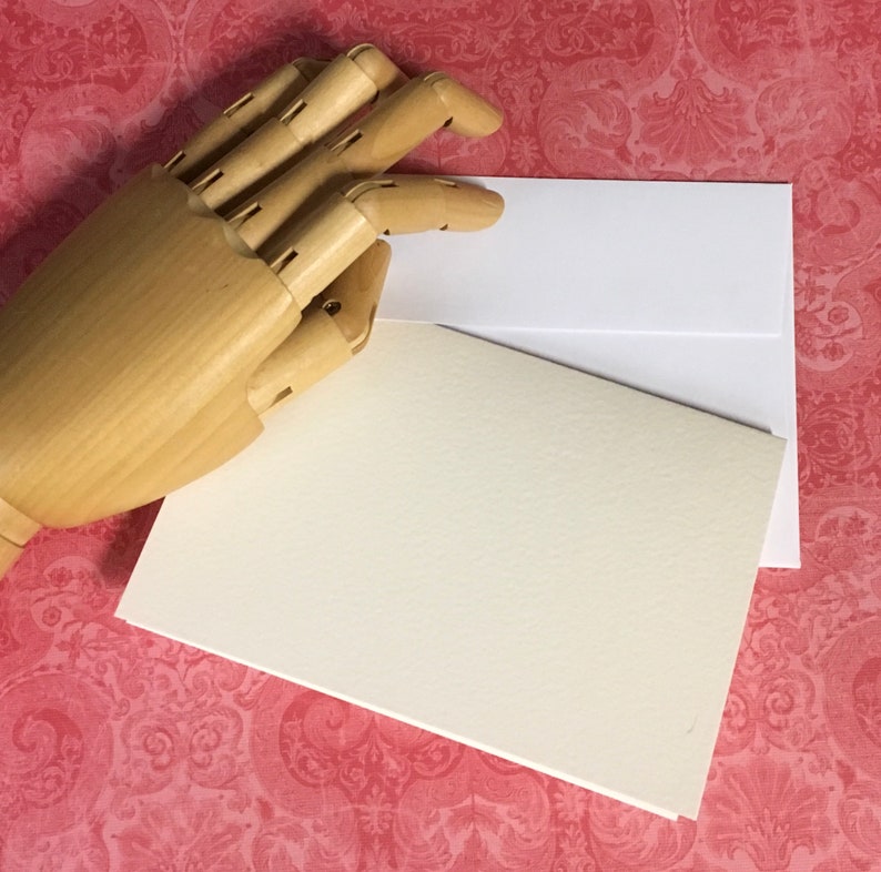 May include: A blank white card with a cream coloured card on top of it. A wooden hand is holding the cards.