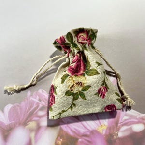 May include: A small, beige drawstring bag with a floral pattern of pink roses and green leaves. The bag is made of a textured fabric and has a natural-colored drawstring closure.