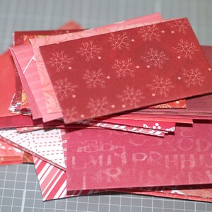 Peut inclure: Une pile de carr&eacute;s de papier rouge &agrave; motifs, avec diff&eacute;rents designs, notamment des flocons de neige et un motif r&eacute;p&eacute;titif de lettres.