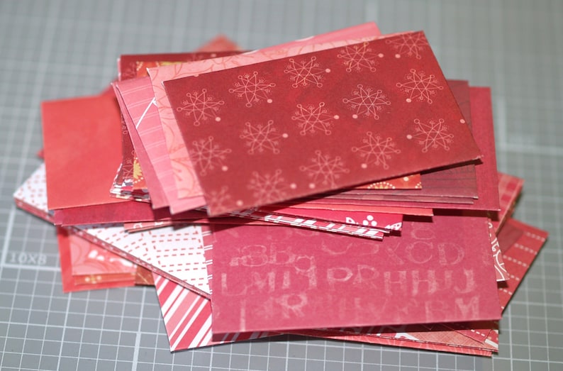 Peut inclure: Une pile de carr&eacute;s de papier rouge &agrave; motifs, avec diff&eacute;rents designs, notamment des flocons de neige et un motif r&eacute;p&eacute;titif de lettres.
