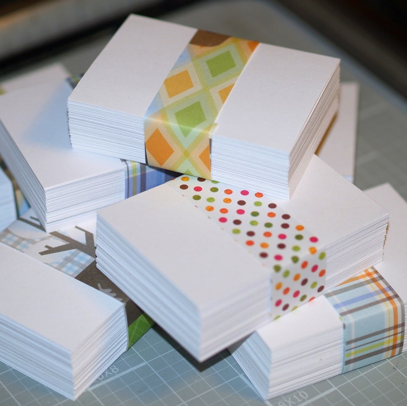 May include: A stack of white note cards with colorful patterned washi tape. The top card has a yellow, green, and white geometric pattern. The second card has a red, orange, and white polka dot pattern. The third card has a blue, white, and green plaid pattern.