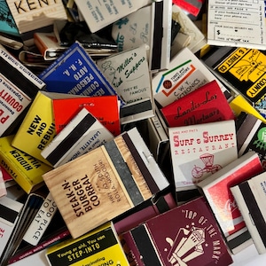 May include: A close-up image of a pile of vintage matchbooks. The matchbooks are colorful and feature various designs and text, including the names of businesses and locations. Some of the matchbooks are red, yellow, blue, and green. The matchbooks are arranged in a random pattern.