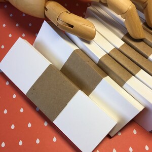 K&ouml;nnte beinhalten: Mehrere Stapel wei&szlig;es Papier, gebunden mit braunen Papierb&auml;ndern, sind diagonal angeordnet. Eine h&ouml;lzerne, bewegliche Hand liegt &uuml;ber dem Papier. Der Hintergrund ist eine orangefarbene Oberfl&auml;che mit wei&szlig;en Tropfenmustern.