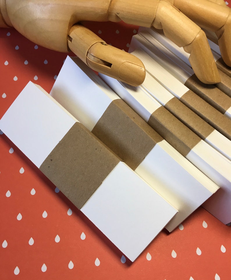 K&ouml;nnte beinhalten: Mehrere Stapel wei&szlig;es Papier, gebunden mit braunen Papierb&auml;ndern, sind diagonal angeordnet. Eine h&ouml;lzerne, bewegliche Hand liegt &uuml;ber dem Papier. Der Hintergrund ist eine orangefarbene Oberfl&auml;che mit wei&szlig;en Tropfenmustern.