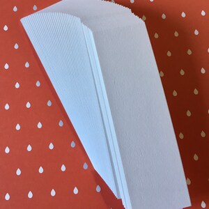 K&ouml;nnte beinhalten: Ein Stapel wei&szlig;er Papiert&uuml;ten mit einem strukturierten, gerippten Design. Die T&uuml;ten sind angewinkelt, wodurch ihre gefalteten Kanten sichtbar werden. Der Hintergrund ist rot-orange mit einem Muster aus wei&szlig;en Tropfen.