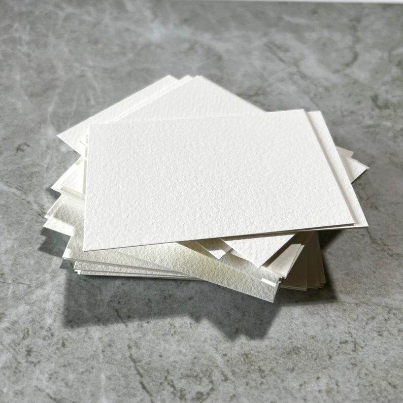 May include: A pile of rectangular, off-white watercolour paper cards with a textured surface. The cards are neatly arranged, with visible edges and corners. The paper appears thick and suitable for painting or drawing.
