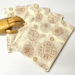 May include: A stack of four cream-colored napkins with a repeating pattern of ornate, golden-brown circular designs. The napkins have a delicate, vintage aesthetic, suitable for decorative use.