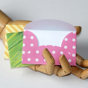 Op de afbeelding: Een roze envelop met witte stippen, opengevouwen om een wit interieur te onthullen. De envelop wordt vastgehouden in een houten hand.