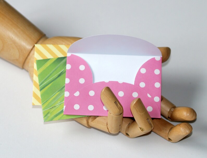 Op de afbeelding: Een roze envelop met witte stippen, opengevouwen om een wit interieur te onthullen. De envelop wordt vastgehouden in een houten hand.