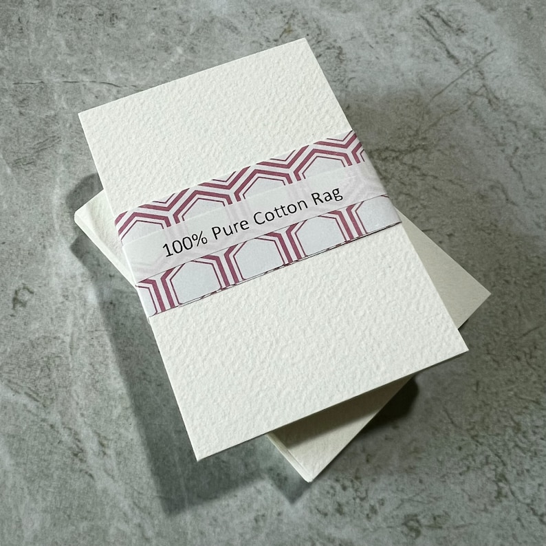 May include: A stack of off-white paper sheets with a textured surface, held together by a decorative band. The band has a burgundy and white geometric pattern, with the text "100% Pure Cotton Rag". The paper is on a grey surface.