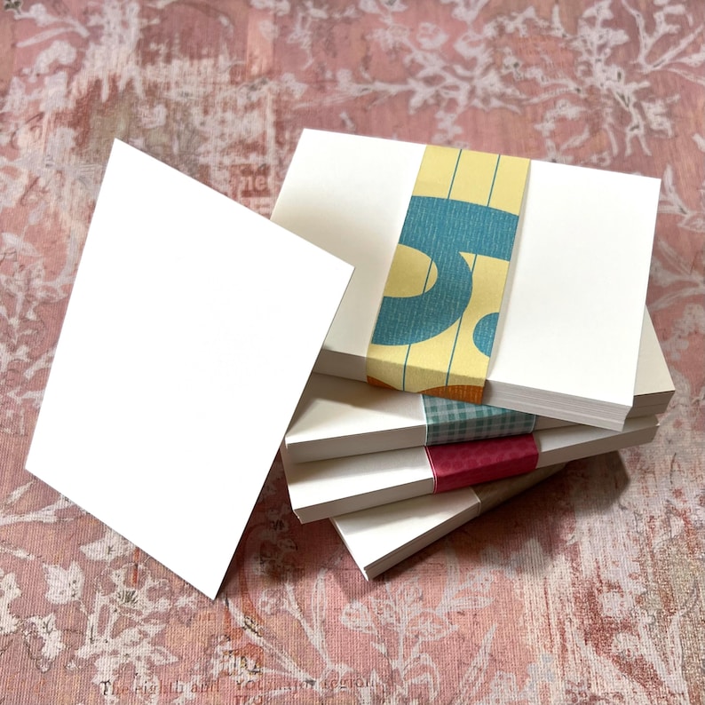 May include: A stack of rectangular white paper cards held together by a decorative paper band featuring a yellow background with blue graphic shapes, followed by a green checkered band and a solid red band with a subtle dot pattern.