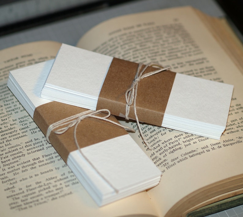 K&ouml;nnte beinhalten: Zwei Stapel blanker wei&szlig;er Papierkarten sind mit Bindfaden zusammengebunden und in braunes Papier eingewickelt. Die Stapel liegen auf einem ge&ouml;ffneten Buch.