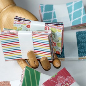 Op de afbeelding: Stapels kleurrijke gepatroneerde vierkante papieren, sommige met strepen, sommige met bloemenpatronen en sommige met geometrische patronen. Het papier is met witte banden bij elkaar gehouden.