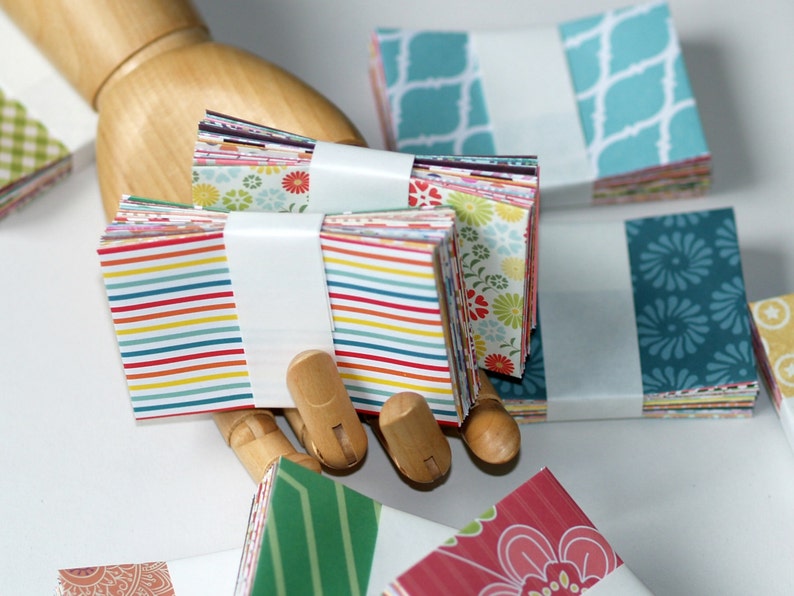 Op de afbeelding: Stapels kleurrijke gepatroneerde vierkante papieren, sommige met strepen, sommige met bloemenpatronen en sommige met geometrische patronen. Het papier is met witte banden bij elkaar gehouden.