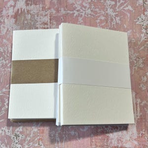 May include: Two stacks of blank white note cards with brown paper backing. The cards have a textured surface.