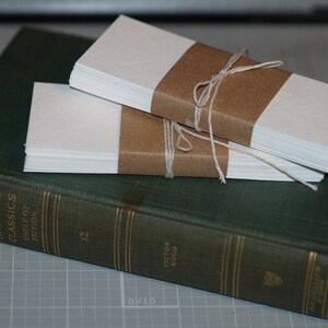 K&ouml;nnte beinhalten: Zwei Stapel blankes wei&szlig;es Papier, jeweils in braunes Papier eingewickelt und mit Schnur gebunden, liegen auf einem gr&uuml;nen Hardcover-Buch. Das Buch tr&auml;gt den Titel "Harvard Classics Shelf of Fiction" und die Nummer "12" auf dem R&uuml;cken. Das Buch liegt auf einer grauen Gitteroberfl&auml;che.