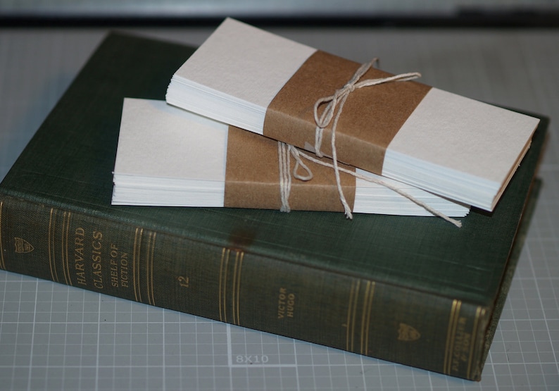 K&ouml;nnte beinhalten: Zwei Stapel blankes wei&szlig;es Papier, jeweils in braunes Papier eingewickelt und mit Schnur gebunden, liegen auf einem gr&uuml;nen Hardcover-Buch. Das Buch tr&auml;gt den Titel "Harvard Classics Shelf of Fiction" und die Nummer "12" auf dem R&uuml;cken. Das Buch liegt auf einer grauen Gitteroberfl&auml;che.