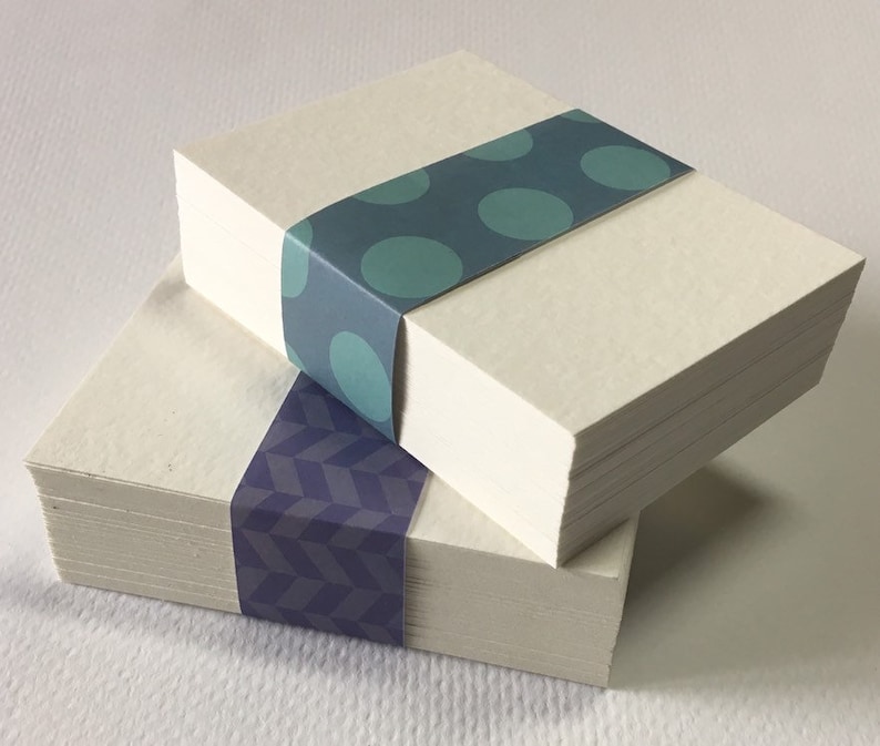 May include: Two stacks of white card stock with patterned paper wrapped around them. The top stack has teal blue polka dots and the bottom stack has a purple geometric pattern.