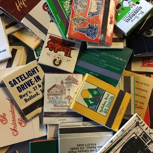 May include: A collection of vintage matchbook covers with various designs and text. Some of the matchbooks feature restaurant names, hotel names, and locations. The matchbooks are in various colors, including red, green, blue, and white.