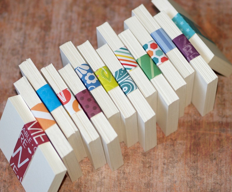 May include: A stack of white note cards with colorful patterned paper bands. The bands feature geometric designs, floral patterns, and abstract shapes.