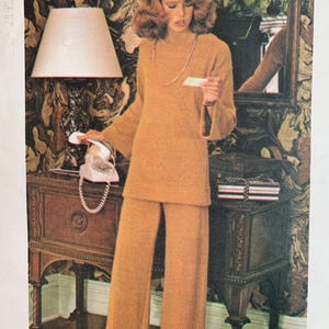 May include: A woman wearing a brown sweater and matching wide-leg pants. She is standing in a room with a patterned wallpaper and holding a phone receiver.