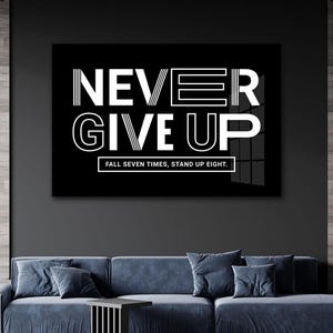 May include: A rectangular wall art piece featuring a black background with the white text NEVER GIVE UP in a bold, stylized font. Below this, a smaller white rectangular outline contains the text FALL SEVEN TIMES, STAND UP EIGHT.