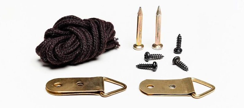May include: A selection of picture hanging hardware, featuring two brass-coloured D-rings, a bundle of dark brown rope, two gold-coloured nails, and four black screws. The items are arranged on a white background.
