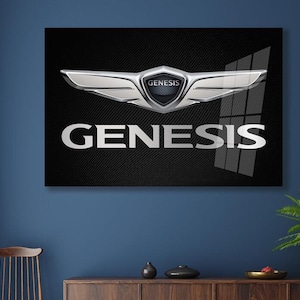 May include: A framed wall art piece featuring the Genesis logo. The logo is a silver emblem with a wing-like design and the word "GENESIS" in silver lettering against a black background. The artwork is displayed on a dark blue wall.