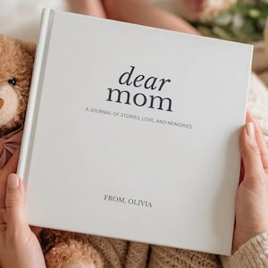 May include: A white journal with the words "dear mom" and "A journal of stories, love, and memories" printed on the cover. A teddy bear with a pink bow and a bouquet of roses are next to the journal. The journal is held by a person.