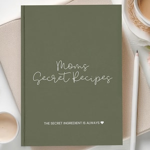 Op de afbeelding: Een rechthoekig olijfgroen receptenboek met witte tekst die Moms Secret Recipes en The Secret Ingredient Is Always leest, gevolgd door een klein hartsymbool. De kaft heeft een matte afwerking en ligt gecentreerd op een lichtbeige textuuroppervlak.
