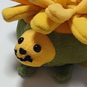May include: A plush toy shaped like a hedgehog with a yellow face and green body. The hedgehog is wearing a yellow flower on its head.