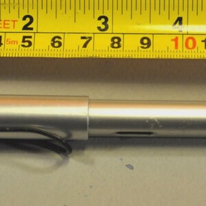 K&ouml;nnte beinhalten: Ein silberfarbener zylindrischer Stift mit schwarzem Clip, der unter einem gelben Ma&szlig;band liegt. Der Stift ist etwa 14 cm lang, was an den sichtbaren Markierungen auf dem direkt dar&uuml;ber liegenden Ma&szlig;band deutlich zu erkennen ist.
