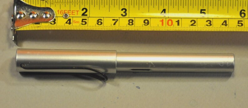 K&ouml;nnte beinhalten: Ein silberfarbener zylindrischer Stift mit schwarzem Clip, der unter einem gelben Ma&szlig;band liegt. Der Stift ist etwa 14 cm lang, was an den sichtbaren Markierungen auf dem direkt dar&uuml;ber liegenden Ma&szlig;band deutlich zu erkennen ist.