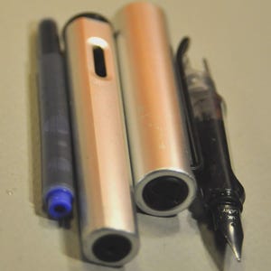K&ouml;nnte beinhalten: Ein zerlegter silberner F&uuml;llfederhalter mit metallischem Finish, bestehend aus einer separaten Tintenpatrone mit blauer Spitze, einem zylindrischen Schaft mit rechteckigem Ausschnitt, einer passenden Kappe und einem schwarzen Griffst&uuml;ck mit einer Metallfeder mit der Aufschrift Lamy.