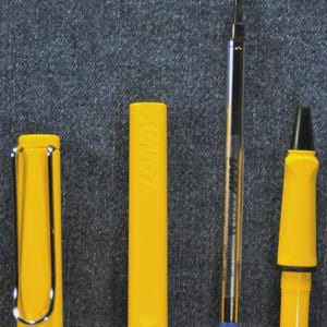 Puede incluir: Un bol&iacute;grafo amarillo desmontado con un clip plateado, un cartucho y una punta. Las piezas del bol&iacute;grafo est&aacute;n dispuestas sobre un fondo de mezclilla. El cartucho tiene el texto "LAMY M63 M" impreso.