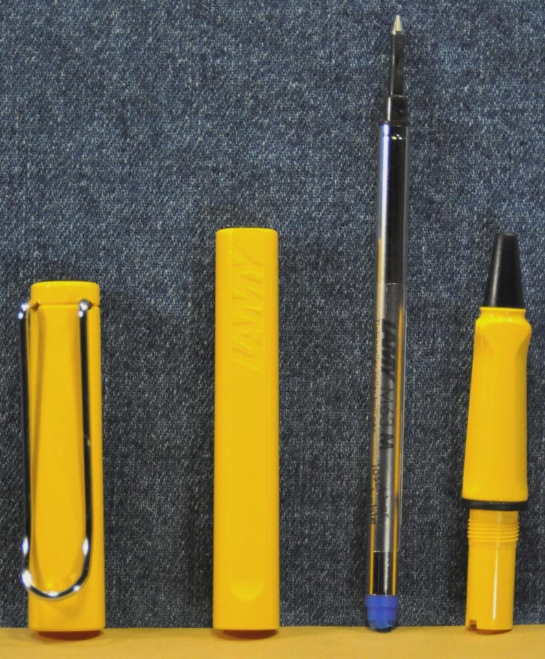 Puede incluir: Un bol&iacute;grafo amarillo desmontado con un clip plateado, un cartucho y una punta. Las piezas del bol&iacute;grafo est&aacute;n dispuestas sobre un fondo de mezclilla. El cartucho tiene el texto "LAMY M63 M" impreso.