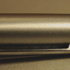 May include: Close-up of a silver-colored pen barrel with a clip. The pen has a brushed metal finish and the word "LAMY" is engraved on the clip. The pen is cylindrical in shape.