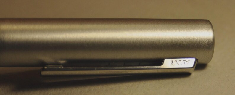 May include: Close-up of a silver-colored pen barrel with a clip. The pen has a brushed metal finish and the word "LAMY" is engraved on the clip. The pen is cylindrical in shape.